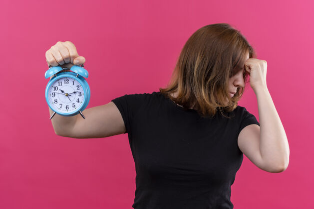 女人紧张的年轻休闲女子拿着闹钟 把拳头放在孤立的粉红色墙上的额头上抱着时钟随意