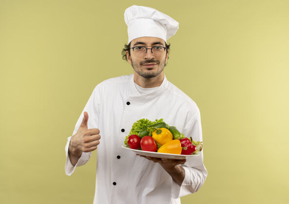 戴着年轻的男厨师穿着厨师制服 戴着眼镜 手里拿着盘子里的蔬菜 他的大拇指孤立地竖立在绿色的墙上盘子眼镜大拇指