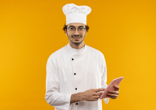 笔记本面带微笑的年轻男厨师穿着厨师制服 戴着眼镜 拿着笔记本孤立在黄色的墙上年轻穿着烹饪