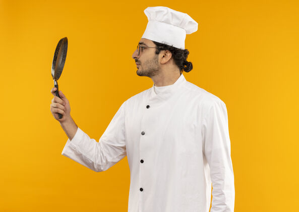 立场站在侧视图年轻的男厨师穿着厨师制服和眼镜拿着和看着煎锅隔离在黄色的墙壁上制服持有薯条