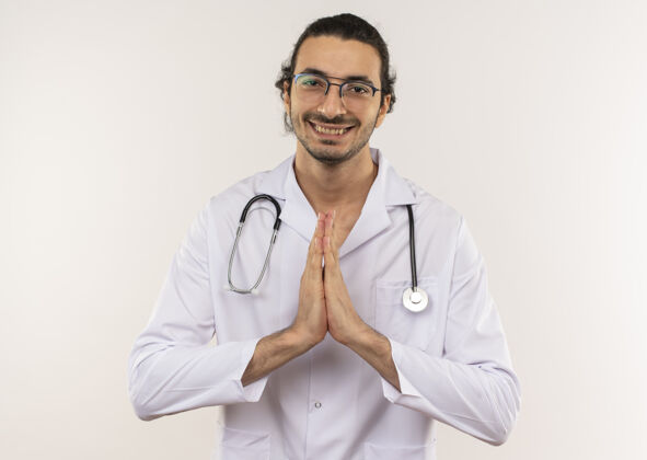 手势微笑的年轻男医生戴着眼镜 穿着白色长袍 听诊器在隔离的白色墙壁上显示祈祷的手势 还有复印空间穿医生年轻