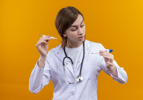 女年轻的女医生穿着医用长袍 戴着听诊器 拿着温度计 看着隔离的橙色墙上的温度计年轻长袍女人