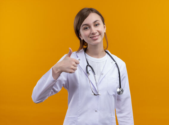 年轻人微笑着的年轻女医生穿着医用长袍和听诊器在孤立的橙色墙上竖起大拇指穿拇指医生