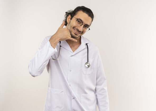 长袍微笑的年轻男性医生 戴着眼镜 身穿白色长袍 听诊器 在隔离的白色墙壁上显示通话手势 并留有复印空间电话眼镜手势