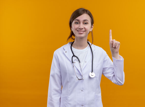 长袍带着微笑的年轻女医生穿着医用长袍和听诊器 在隔离的橙色墙壁上竖起手指 留着复印空间听诊器女人医生