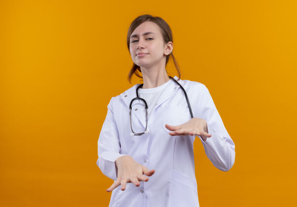 伸展穿着医用长袍和听诊器的年轻女医生在隔离的橙色墙壁上伸展双手 并留有复印空间穿女人医学