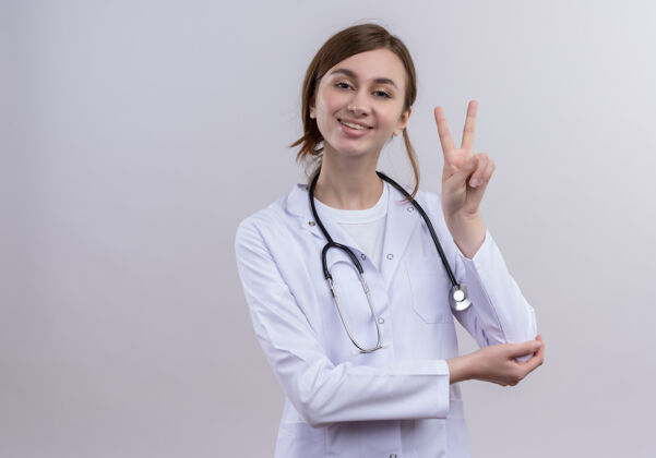 医学带着微笑的年轻女医生穿着医用长袍 听诊器在隔离的白墙上做着和平的手势 还有复印空间听诊器女性穿
