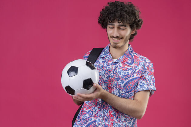 微笑微笑着的年轻英俊的卷发男子拿着足球 看着它在孤立的粉红色墙上复制空间足球卷发球