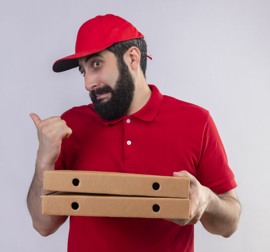 制服印象深刻的年轻英俊的白人送货员穿着红色制服 戴着帽子拿着披萨盒 指着身后孤立的白色帽子盒子印象