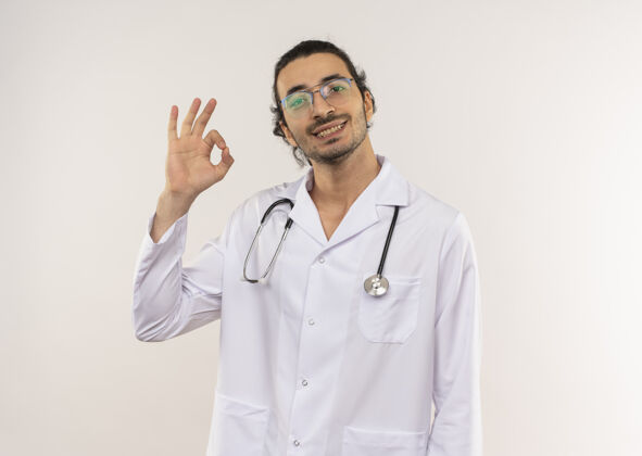 微笑微笑的年轻男性医生 戴着眼镜 穿着白色长袍 听诊器 在隔离的白色墙壁上显示出良好的姿势 并留有复印空间听诊器年轻展示
