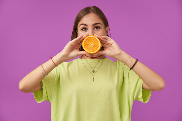 休闲一个漂亮漂亮的女孩的肖像 她用两只手把橘子捂在嘴上 站在紫色的墙上 穿着绿色的t恤 手镯 戒指和项链T恤女孩热带
