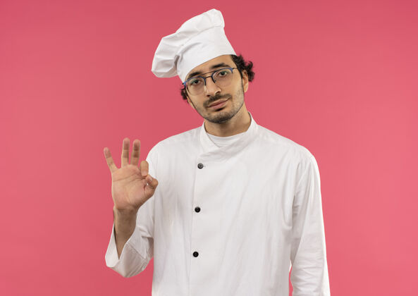 年轻年轻的男厨师穿着厨师制服 戴着一副眼镜 在粉红色的墙上显示出很好的姿势男性眼镜制服