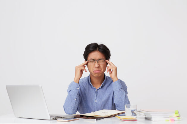 压力一个戴着眼镜 眼睛闭着摸太阳穴 心情紧张 心情不愉快的亚洲年轻商人 在白墙上的桌子旁用笔记本电脑感到压力封闭互联网笔记本电脑
