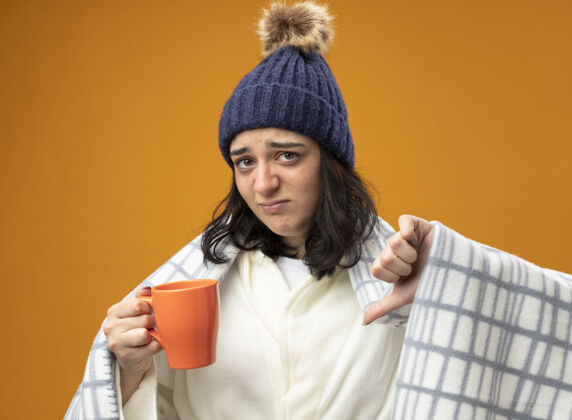 茶年轻的白种人生病的女孩穿着长袍 戴着格子布的冬帽 手里拿着一杯茶 看着相机 拇指朝下 孤立地站在橙色的背景上相机格子杯子