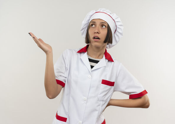 烹饪印象深刻的年轻女厨师穿着厨师制服 一边看一边显示空手 把手放在腰部隔离在白色年轻显示印象