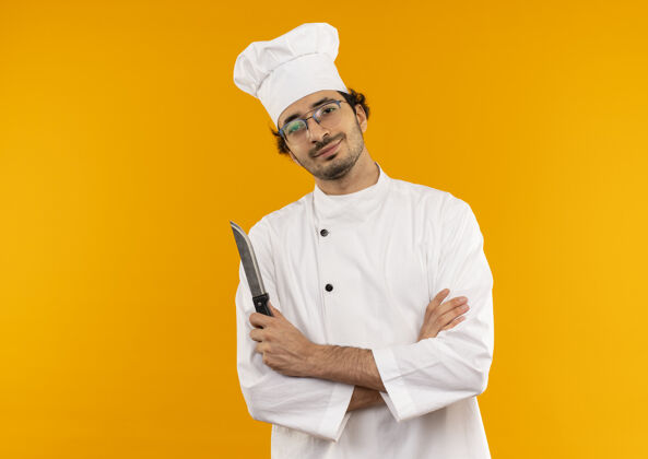 制服高兴的年轻男厨师穿着厨师制服和眼镜拿着刀和交叉的手隔离在黄色的墙上眼镜刀烹饪