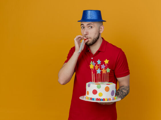 相机令人印象深刻的年轻英俊的斯拉夫党的家伙戴着党的帽子看着相机拿着生日蛋糕与党吹嘴隔离在橙色背景与复制空间帅哥蛋糕小伙子