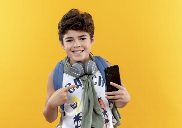 小面带微笑的小男孩背着书包 戴着耳机 指着隔离在黄色墙上的电话耳机包微笑