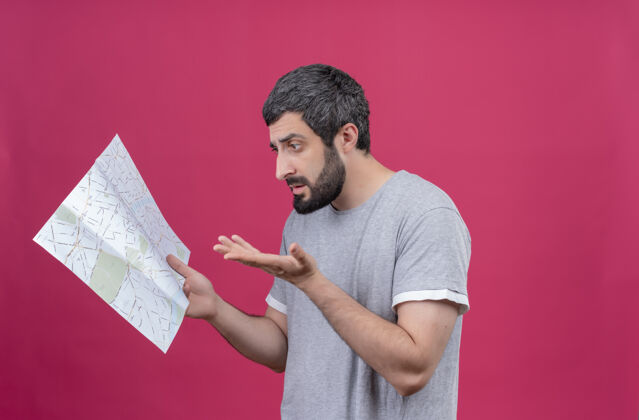 持有困惑的年轻英俊的白人旅行家男子拿着地图 看着地图 显示空的手孤立在粉红色的复制空间空间粉色旅行者