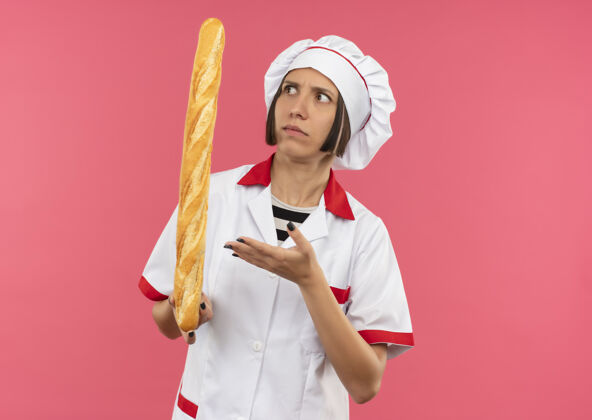女性困惑的年轻女厨师身着厨师制服 手拿面包棒看着并指着孤立的粉红色的复印空间坚持年轻持有