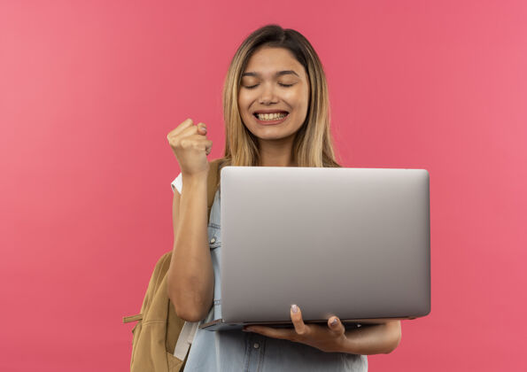 拳头快乐的年轻漂亮的女学生 背着包拿着笔记本 紧握拳头闭着眼睛 隔离在粉色的空间里笔记本电脑穿握紧