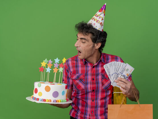 钱中年白种人聚会男子戴着生日帽拿着生日蛋糕纸袋礼包和钱看着蛋糕准备咬它绿色背景隔离举行准备帽子