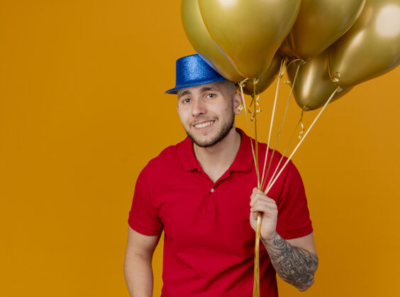 年轻面带微笑的年轻英俊的斯拉夫党人戴着党的帽子拿着气球看着相机孤立的橙色背景与复制空间帽子微笑相机