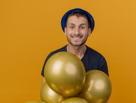 气球面带微笑的白人帅哥戴着蓝色的派对帽 手里拿着氦气球 这些气球被隔离在橙色的背景上 还有复制空间举行帽子微笑