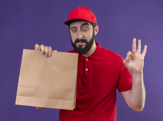 纸印象深刻的年轻英俊的白种人送货员身穿红色制服 戴着帽子 看着纸包 做着“行”字孤立在紫色上Ok男人穿着