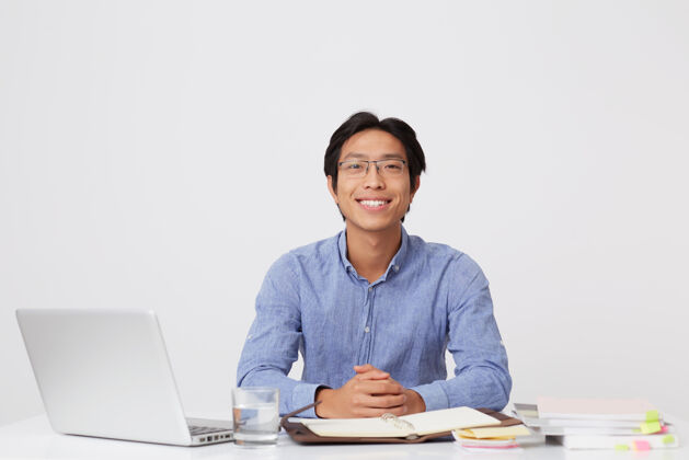 坐着的人快乐英俊的亚洲年轻商人戴着眼镜 穿着蓝色衬衫 坐在桌边 笔记本电脑盖在白墙上高兴学生眼镜