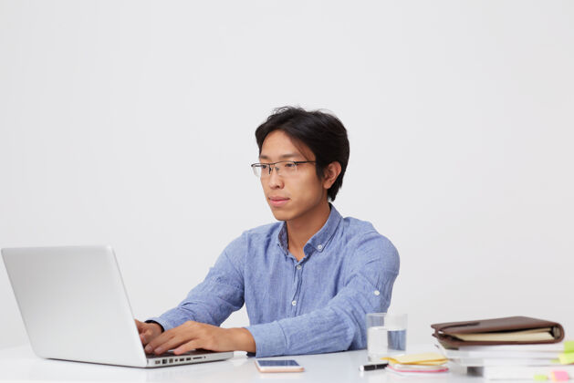 专家严肃专注的亚洲年轻商人戴着眼镜坐在桌子旁打字 工作时手提电脑隔着白墙工作场所笔记本电脑沉思