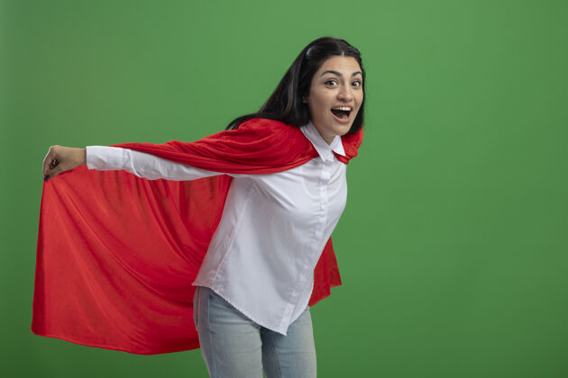 相机顽皮的年轻白种人超级英雄女孩拿着她的英雄斗篷 代表飞行 看着相机在绿色背景与复制空间隔离年轻女孩超级英雄