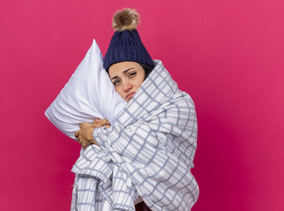 围巾虚弱的年轻白种人生病的女孩戴着冬天的帽子和围巾裹在格子布抱枕上把头放在上面看着摄像机在深红色的背景下与复制空间隔离头包裹相机