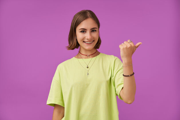 女士漂亮漂亮的女孩的肖像微笑着用拇指指着紫色墙上的复制空间穿着绿色t恤 牙套 项链和手镯女士牙齿项链