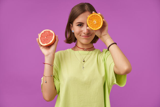 休闲有魅力的可爱女孩的肖像 深色短发用橙色遮住眼睛 遮住一只眼睛站在紫色的墙上穿着绿色t恤 项链 背带和手镯女士背带女孩
