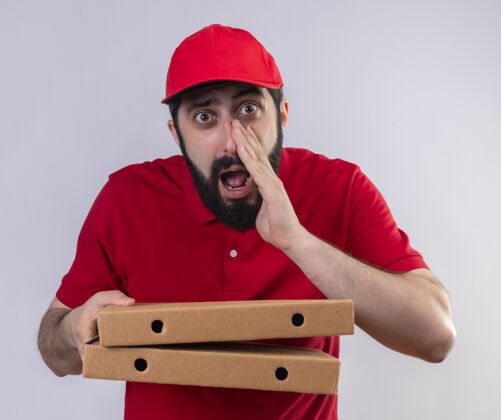 披萨印象深刻的年轻英俊的白人送货员穿着红色制服 戴着帽子拿着比萨饼盒 在白色的地板上低语拿着送货帽子