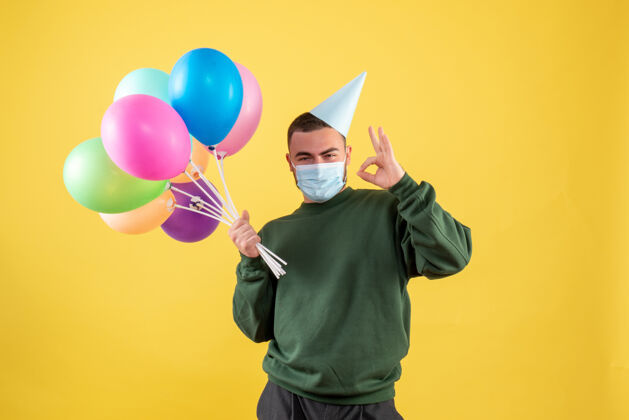 背景正面图黄色背景上手持彩色气球的年轻男性快乐气球年