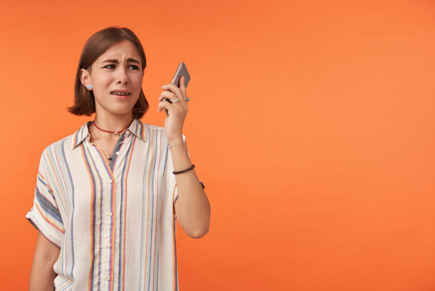 悲伤不开心的女孩拿着智能手机看穿着条纹衬衫 戴着牙套和手镯看着右边 在橙色的墙上复制空间时尚电话牙齿