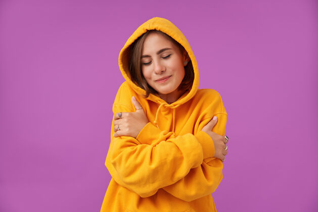 女性女学生 一头浅棕色短发的年轻女士 拥抱着自己 感觉温暖舒适 闭上眼睛站在紫色的墙上穿着橙色的连帽衫和戒指温暖穿孔拥抱