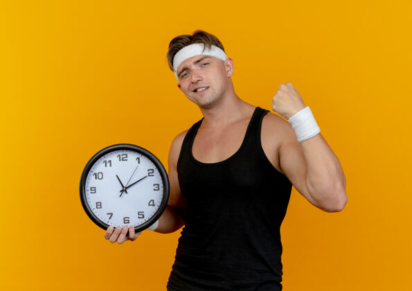 握紧自信的年轻帅气的运动型男人戴着头带和腕带拿着时钟和握紧拳头孤立在橙色自信年轻头带