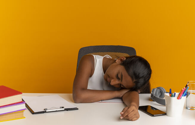 书桌疲惫的年轻女学生坐在课桌旁 把学习工具放在桌子上 孤立地放在橙色的墙上累了坐着年轻