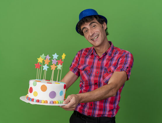 中年快乐的中年白种人党男子戴着党的帽子伸展生日蛋糕看着相机隔离在绿色的背景帽子欢乐蛋糕