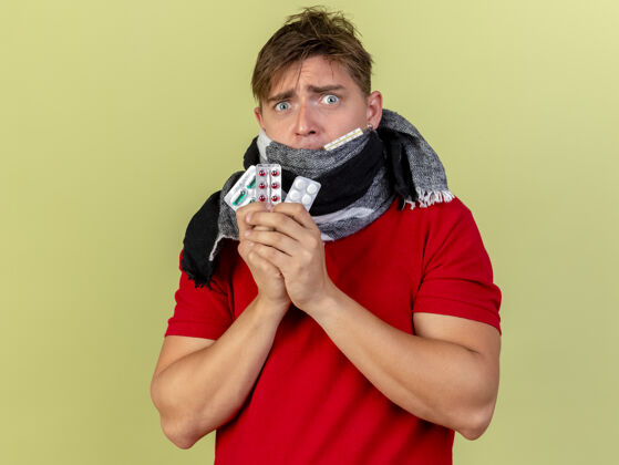 金发印象深刻的年轻英俊的金发病男子戴着围巾 嘴里拿着温度计和药片包 看着橄榄绿背景上的相机与复制空间隔离温度计围巾橄榄
