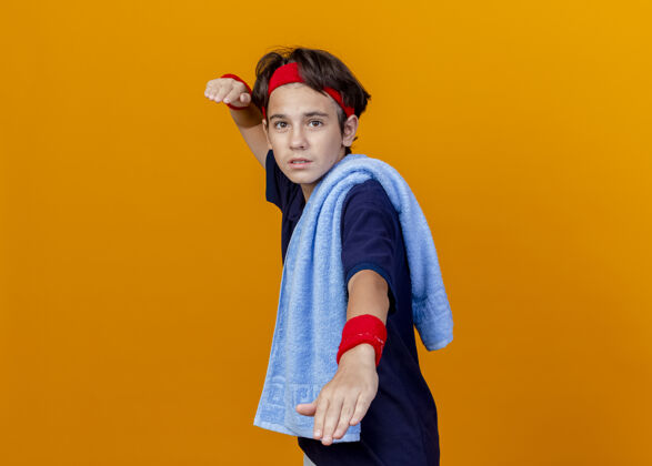 运动年轻帅气的运动男孩戴着头带和护腕 肩上戴着牙套和毛巾 站在侧视图中 与橙色背景隔离 留有复制空间年轻腕带肩膀