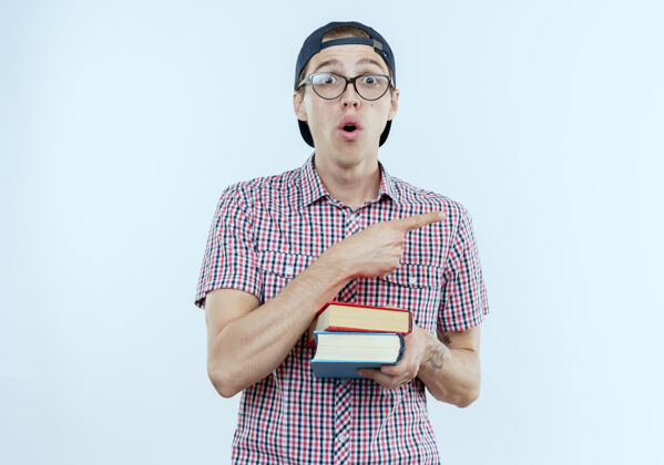 眼镜惊讶的年轻学生男孩戴着书包 眼镜和帽子拿着书 指着隔离在白色墙壁上的复印空间男孩壁板包