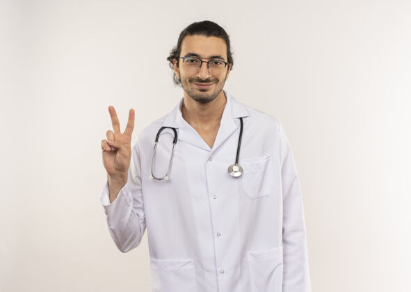 年轻年轻的男医生戴着眼镜 身穿白色长袍 听诊器 在隔离的白色墙壁上显示和平的姿态 并留有复印空间和平手势显示