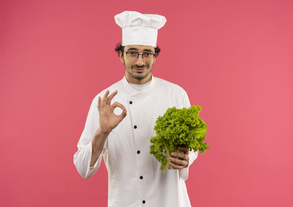 厨师年轻的男厨师穿着厨师制服 戴着眼镜 手里拿着色拉 在粉红色的墙上显示出很好的姿势年轻烹饪表演