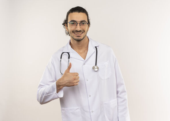 穿微笑着的年轻男医生 戴着眼镜 穿着白色长袍 手持听诊器 他的大拇指竖立在与世隔绝的白色墙上 留着复印空间长袍年轻眼镜