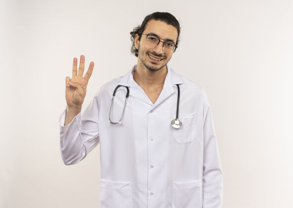 光学微笑的年轻男性医生 戴着眼镜 穿着白色长袍 听诊器 在隔离的白墙上显示三个 还有复印空间眼镜微笑年轻