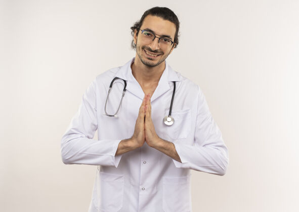 光学微笑的年轻男医生戴着眼镜 穿着白色长袍 听诊器在隔离的白色墙壁上显示祈祷的手势 还有复印空间眼镜男性手势